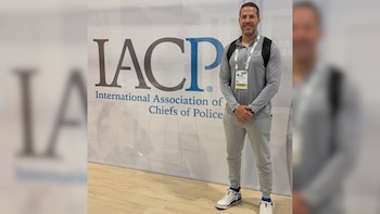 Edwin Lopez, con camisa gris y pantalones deportivos, de pie sonriendo frente a un gran cartel blanco y azul de la Asociación Internacional de Jefes de Policía