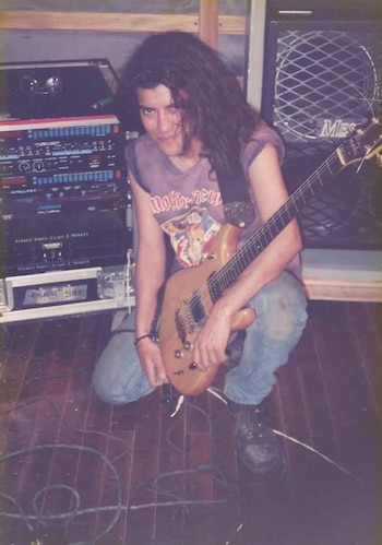 Hombre de cabello oscuro y largo, con remera sin mangas, jeans y botas, sostiene una guitarra eléctrica. Hay equipos de audio y un amplificador