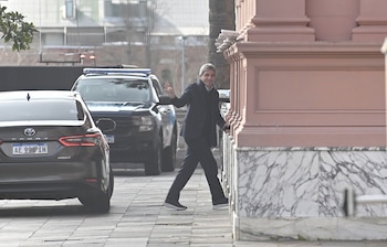 Un hombre con traje oscuro saluda con la mano mientras camina junto a un edificio rosado con automóviles oficiales detrás