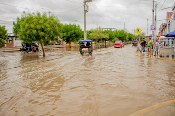 Calle cubierta de agua turbia por donde circulan mototaxis y un automóvil rojo frente a peatones parados sobre la acera