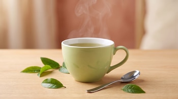 Taza verde con té caliente humeante, hojas de té verdes y frescas, una cuchara de metal plateada y mesa de madera clara con fondo difuminado.