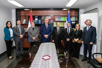 Ocho personas, cinco hombres y tres mujeres, posan formalmente frente a una mesa larga, un estante de libros y banderas en una oficina