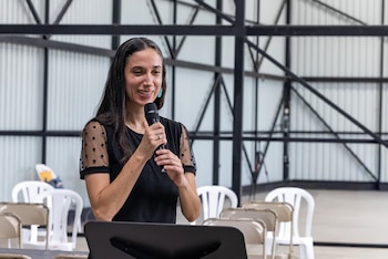 Una mujer sonriente con cabello oscuro y blusa negra con mangas de lunares transparentes sostiene un micrófono frente a un atril, con sillas vacías y vigas metálicas de fondo