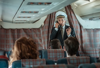 Un hombre con uniforme de piloto habla por teléfono dentro de un avión frente a pasajeros sentados de espaldas