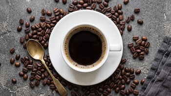 Vista superior de una taza con café negro, granos de café tostados dispersos, una cuchara dorada y un paño gris sobre piedra