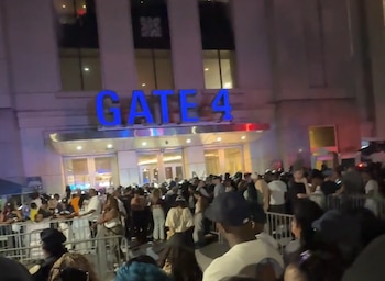 Vista nocturna de una multitud de personas frente a la entrada de un edificio con el letrero luminoso azul 'GATE 4'. Hay vallas metálicas