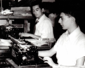Imagen en blanco y negro de dos hombres jóvenes sentados en escritorios, tecleando en máquinas de escribir antiguas
