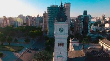 Vista aérea de una torre de reloj blanca con cúpula oscura, rodeada de edificios urbanos y áreas verdes, bajo un cielo despejado
