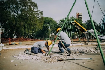 Dos obreros con cascos amarillos reparan la base de una estructura verde de juegos infantiles. Hay escombros de hormigón en el suelo y árboles al fondo