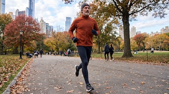 Hombre corriendo con gorro, chaqueta naranja, pantalones oscuros y guantes en un parque con árboles de hojas otoñales y rascacielos al fondo.