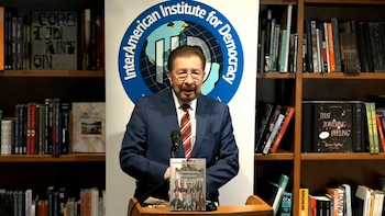 Hombre con traje y corbata ante un atril con micrófono, sosteniendo un libro. Fondo con un logotipo circular azul y estanterías llenas de libros