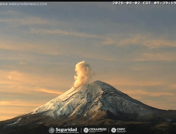 Un volcán cubierto de nieve con una columna de humo y ceniza contra un cielo con tonos de amanecer. Texto de fecha y hora, y logotipos de instituciones