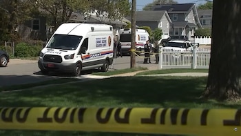 Furgonetas de la Unidad de Búsqueda de Escena del Crimen de Nassau County y policías en una calle residencial, con cinta amarilla visible. Casas y árboles en el fondo