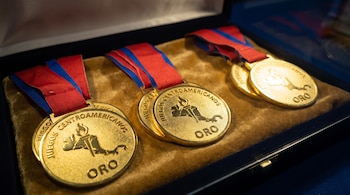 Tres medallas de oro con cintas rojas y azules en un estuche. Las medallas tienen el texto 'Juegos Centroamericanos' y 'Oro' junto a un mapa con una antorcha.