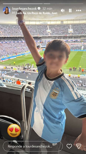 Niño con el rostro difuminado y la camiseta argentina levanta la mano en la tribuna de un estadio de fútbol.
