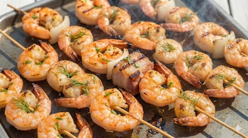 Brochetas de langostinos y panceta cocinándose sobre una parrilla metálica, mostrando un dorado uniforme y un sutil humo