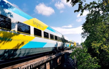 Tren de pasajeros Brightline con colores blanco, azul y amarillo en las vías férreas, árboles verdes, cielo azul con nubes blancas