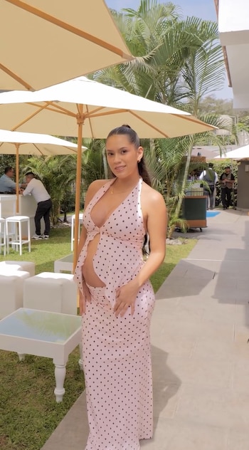 Mujer embarazada con vestido largo rosa de lunares negros. Entorno exterior con sombrillas color crema, mesas y sillones blancos, plantas y palmeras de fondo