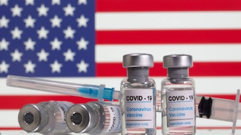 Múltiples viales de la vacuna contra el COVID-19 con etiquetas "Coronavirus Vaccine" y jeringuillas sobre un fondo borroso de la bandera de los Estados Unidos