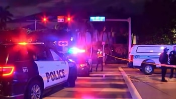 SUV policial con luces de emergencia rojas y azules en calle nocturna. Otros vehículos, agentes y cinta amarilla de seguridad visible en la escena