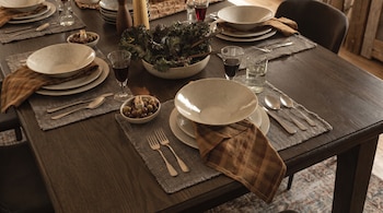 Mesa de madera oscura con seis puestos, cada uno con plato, cubiertos, copa y servilleta. Hay una ensaladera con hojas verdes y cuencos con aceitunas.