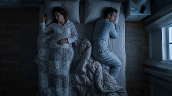 Vista cenital de una cama doble. Mujer duerme boca arriba, hombre boca abajo, ambos con pijama y cubiertos, en una habitación oscura por la noche.