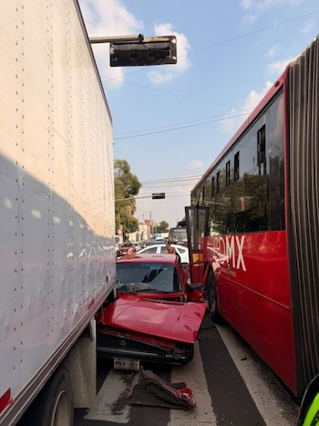 Un automóvil rojo chocado y deformado se encuentra atrapado entre la caja blanca de un camión y el costado de un autobús articulado rojo