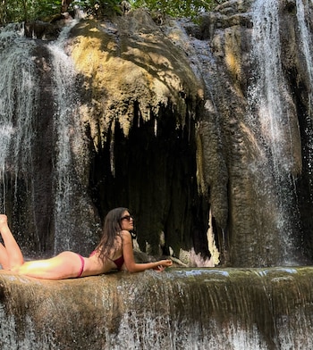 Una mujer en bikini recostada sobre una formación rocosa al pie de una cascada natural