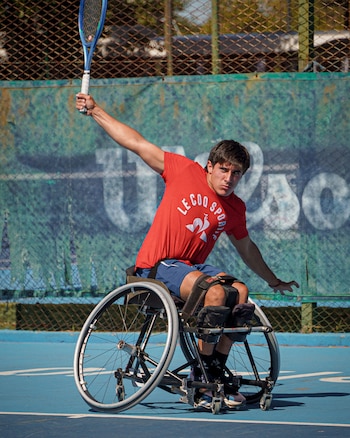 Un hombre joven, Gonzalo Lazarte, en una silla de ruedas deportiva en una cancha de tenis azul, levanta una raqueta azul para golpear una pelota