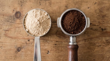 Cuchara medidora con proteína en polvo beige y portafiltro de café molido oscuro sobre una superficie de madera clara, con nudos visibles.