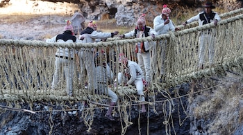 Seis hombres con gorros de lana teñidos y trajes claros ajustan las cuerdas de un puente de fibra vegetal tendido sobre un barranco de rocas.