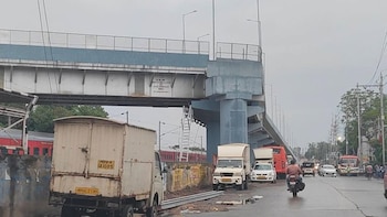 Puente ferroviario de cemento y metal con giro de 90 grados sobre una calle con vehículos, un tren rojo en vías inferiores y personas