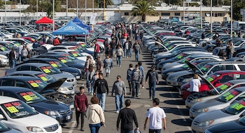 Una multitud de personas camina entre largas filas de autos usados estacionados en un gran mercado al aire libre, con carteles de venta y capós abiertos.