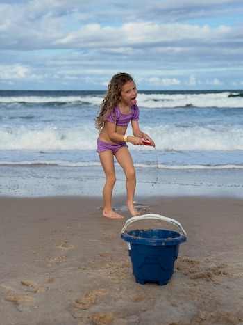 Una niña con cabello rizado y bañador morado juega en la arena de la playa con una pala roja y un balde azul. El mar y el cielo nublado están de fondo