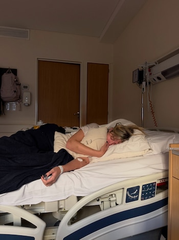 Mujer rubia acostada en cama de clínica, cubierta con manta oscura. Tiene una pulsera identificatoria y una vía intravenosa en su brazo. Mochila y aparatos médicos en la pared
