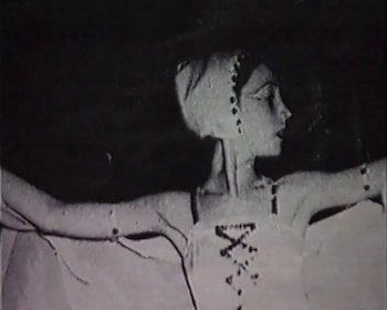 Imagen en blanco y negro de una mujer de perfil con los brazos extendidos, ataviada con tocado con perlas y vestido con corsé sobre un fondo oscuro