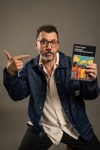 Un hombre con anteojos y chaqueta de mezclilla sostiene un libro con una mano y lo señala con el dedo de la otra mano