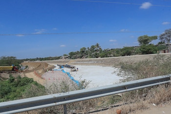 Sitio de construcción de obras de prevención con lecho de río, trabajadores, maquinaria, tuberías, concreto, guardarraíl y vegetación bajo cielo azul