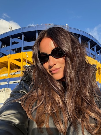 Selfie de una mujer con cabello castaño largo y gafas de sol, sonriendo frente a la estructura azul y amarilla del estadio La Bombonera bajo el cielo azul