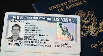 Primer plano de una visa B2 de turista de Estados Unidos con foto, datos personales y la Estatua de la Libertad, y un pasaporte azul oscuro al fondo.
