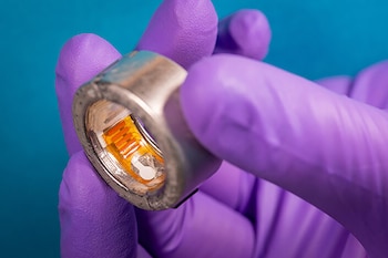 Una mano con guante violeta sostiene un anillo metálico con circuitos electrónicos anaranjados y un sensor blanco en su interior