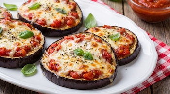 Rodajas de berenjena horneadas cubiertas con salsa de tomate, queso fundido, hierbas y albahaca en un plato. Un cuenco con salsa y un mantel a cuadros rojos.