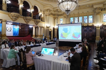 Vista general de una sala de reuniones con personas sentadas en mesas, un proyector mostrando mapas de Guatemala y banderas en el fondo