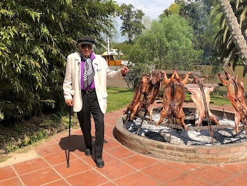 Hombre mayor, Julio Le Parc, con sombrero negro, gafas de sol y bastón, de pie frente a un asado con corderos enteros al fuego, rodeado de vegetación y un río con un bote blanco