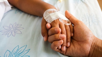 Primer plano de una mano adulta que sostiene la mano de un niño, quien tiene un vendaje y una vía intravenosa en la muñeca. El fondo es una sábana clara con patrones.