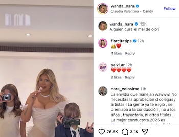 Wanda Nara, de cabello rubio, en un vestido blanco escotado, sostiene un chupete rosa. A sus lados, dos niñas con cámaras de fotos y video