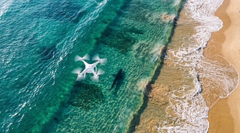 Vista aérea de un dron blanco sobre el agua turquesa cerca de la silueta de un tiburón y la playa.