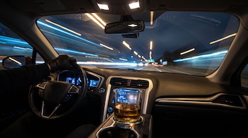 Un vaso con líquido ámbar en la consola de un vehículo, una mano con guante al volante y luces de velocidad en el parabrisas.