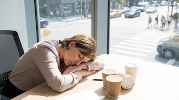 Mujer con camiseta de color claro dormida en un escritorio de madera con vasos de café desechables y un bolígrafo, junto a una ventana con vista urbana.