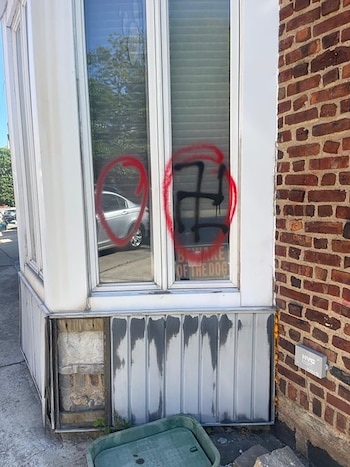 Ventanas de un edificio con grafitis de una esvástica negra y un círculo rojo; se ve un cartel de 'Cuidado con el perro' y parte de una pared de ladrillo